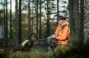 Bussföraren Göran i jaktkläder sittandes i huk i skogen tillsammans med sin jakthund.