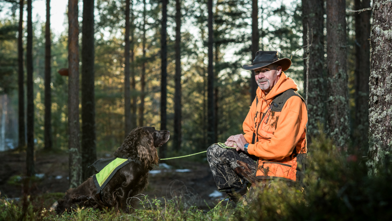Bussföraren Göran i jaktkläder sittandes i huk i skogen tillsammans med sin jakthund.